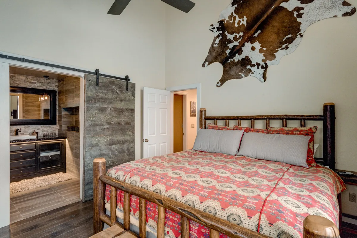 A spacious bedroom featuring a large wooden bed dressed in cozy bedding, accented by cowhide wall art above the headboard. The room connects to an en suite bathroom with dark cabinetry, visible through a partially open doorway, suggesting ensuite convenience and privacy.