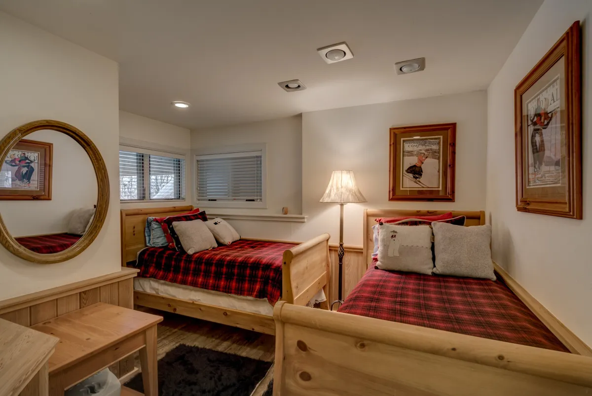 A warm, inviting bedroom with two twin beds clad in plaid bedding, a decorative mirror above, and playful artwork on the walls. Soft lighting and a side table encourage restful nights or cozy gatherings for kids or friends.