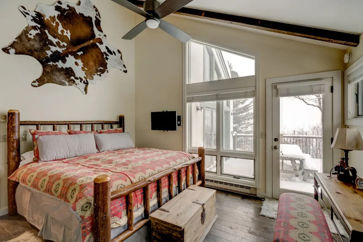 This image features a cozy bedroom with patterned bedding, cowhide wall art, and large windows that open onto a snowy landscape. Wooden furniture adds to the inviting atmosphere, and the overall feel is of a warm retreat with beautiful outdoor views.