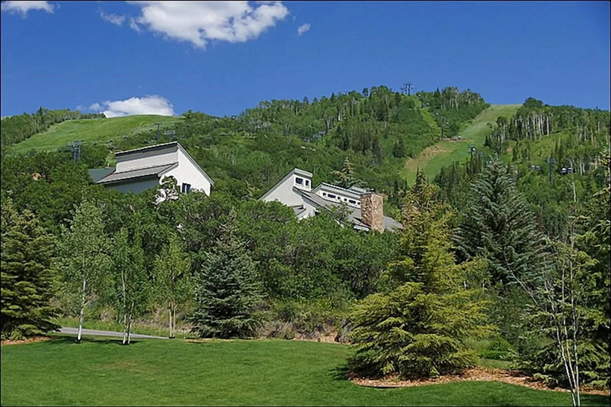 The hero image showcases the town house nestled against a serene, sloping hillside, surrounded by mature trees under a brilliant blue sky. The setting feels tranquil and private, with beautiful sunrise light washing over the landscape and hints of outdoor adventure all around.