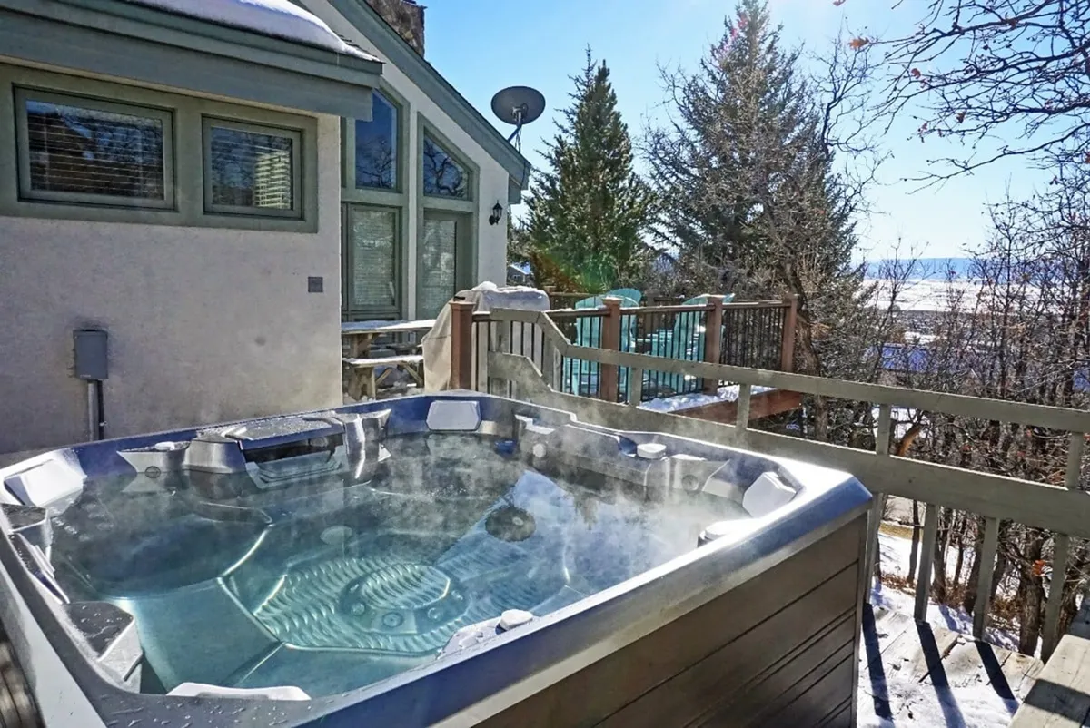 An exterior image showcasing the chalet’s inviting hot tub on a wooden deck, surrounded by tall trees and the house’s large windows. The setting is tranquil, with ample space for groups to gather, socialize, or unwind while enjoying nature and the fresh mountain air.
