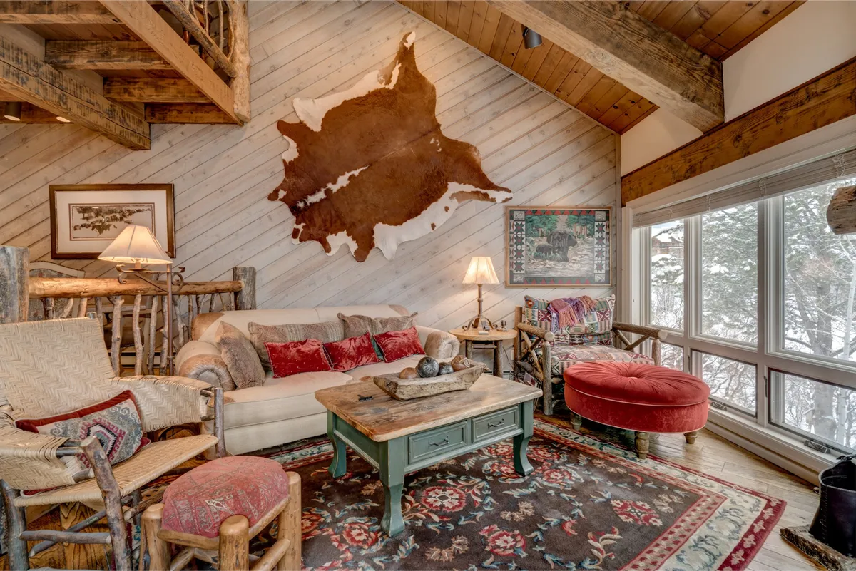 A spacious living room with a comfortable sofa, coffee table, and area rug. Large windows let in abundant natural light and display snowy outdoor views. Cowhide wall art and inviting decor create a cozy, welcoming atmosphere, ideal for both group gatherings and intimate conversations.