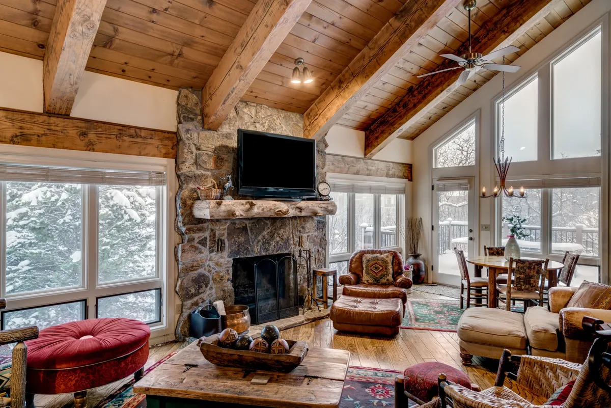 A stunning interior with a stone fireplace, large windows framing snowy mountain views, and rustic furnishings. The scene exudes mountain retreat energy—perfect for watching snowfall after skiing or drying off gear by the fire.