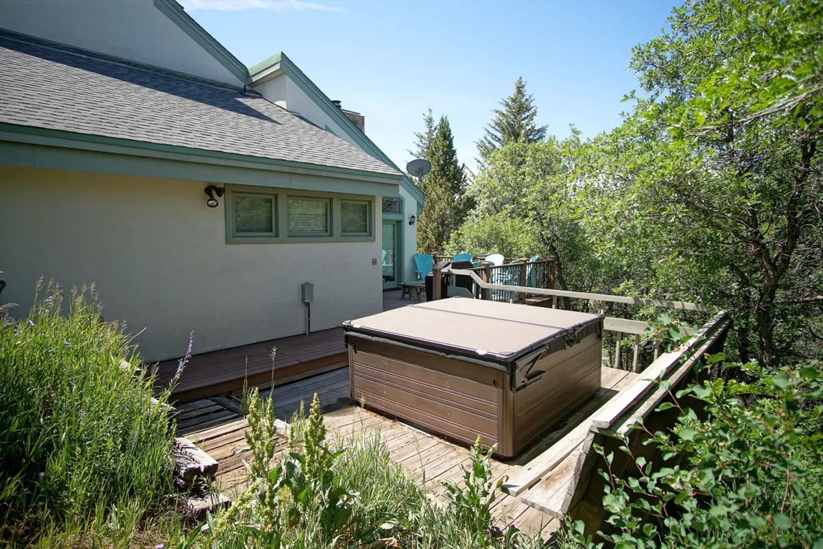 An inviting exterior scene featuring a wooden deck surrounded by greenery. The main focal point is a hot tub, with ample space around it for relaxation and socializing. The deck is well-integrated into the natural landscape, offering a peaceful spot to enjoy the outdoors.