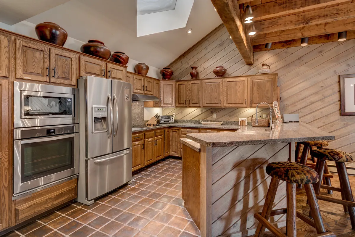 A modern kitchen featuring stainless steel appliances, granite countertops, wooden cabinets, and stylish bar stools. The space is designed for both culinary creativity and socializing, with decorative pottery adding warmth and character.